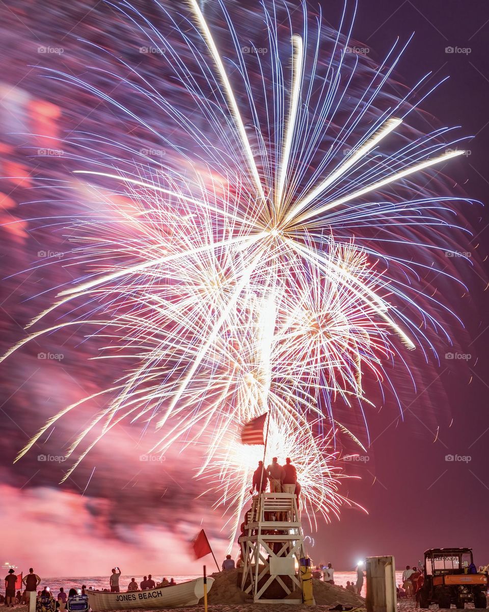Fireworks celebration with people standing on a lifeguard tower on the beach with an American flag