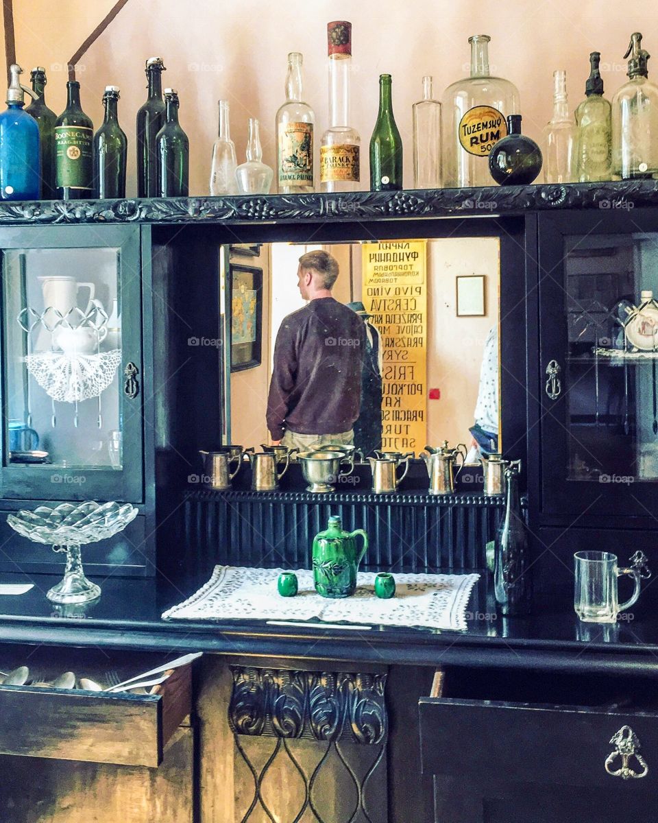 Reflection in the mirror of man standing in a room full of old antique bottles and glasses. 