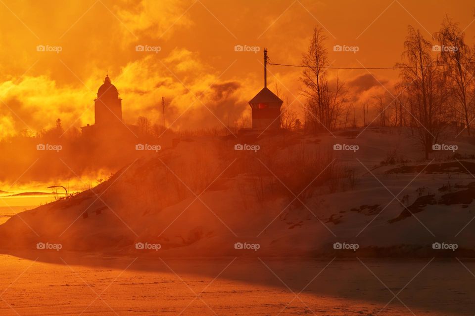 Blazing morning sunrise behind small beacon or lighthouse on the rocky island in front of Suomenlinna fortress island on an extremely cold winter morning at sunrise with sea fog and icy Baltic sea, the Suomenlinna church.