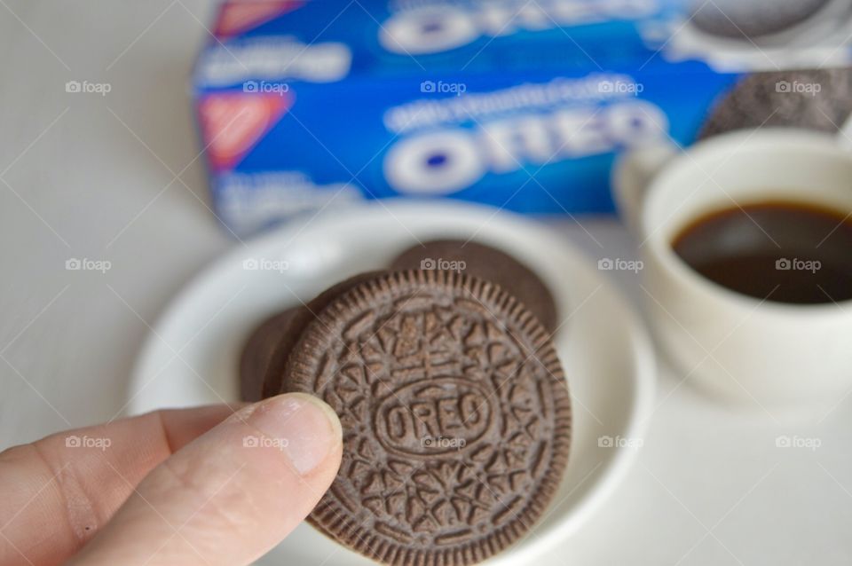 A persons fingers holding an Oreo cookie and a cup of coffee in the background 