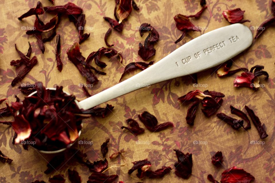 Overhead view of a stainless steel measuring spoon imprinted with the phrase, “1 CUP OF PERFECT TEA,” filled with dried hibiscus petals on a brown and burgundy patterned surface with scattered hibiscus tea 