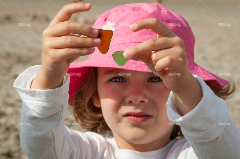 Little girl in pink hat holding up two stained pieces of smooth shaped glass.