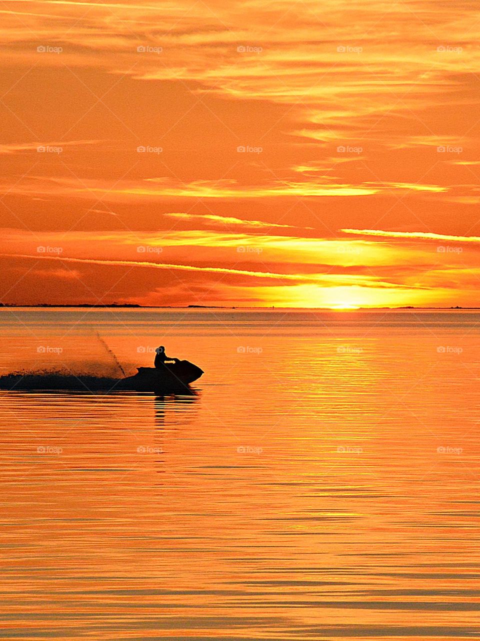 A silhouette of a man and his skidoo crosses the bay during a descending sunset as it throws up a rooster tail