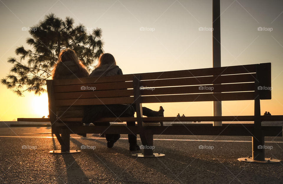 Two Girls Sitting On The Bench Outside And Enjoying The Sunset
