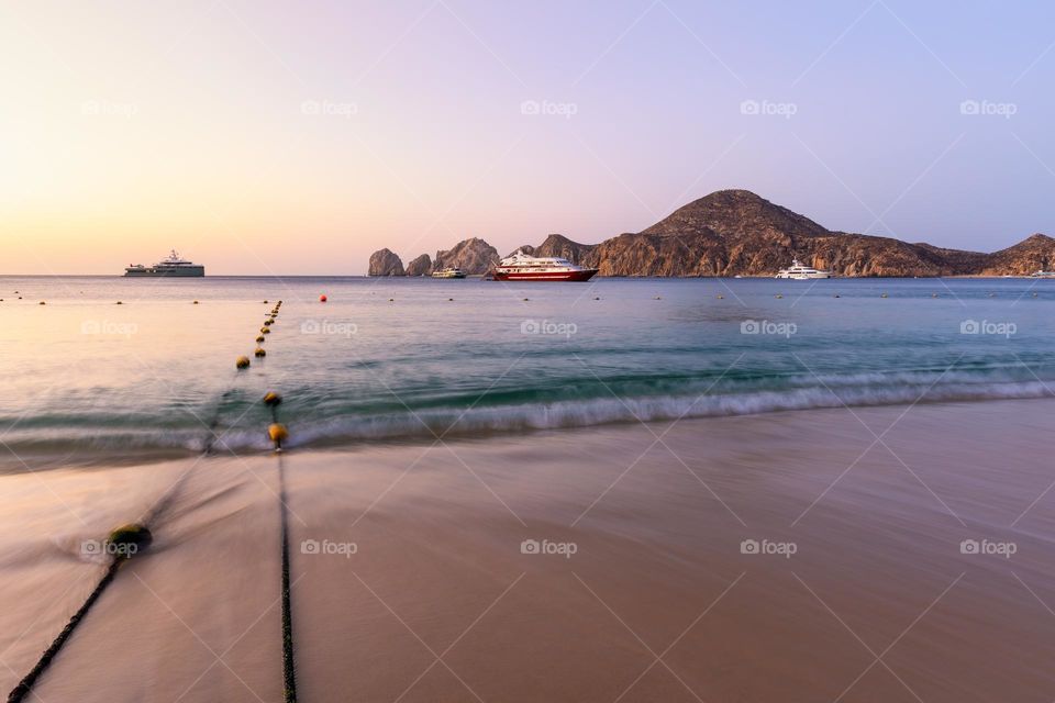 Waves crashing on a beach at sunrise, with a boat in the ocean in front of a mountain and rock formations