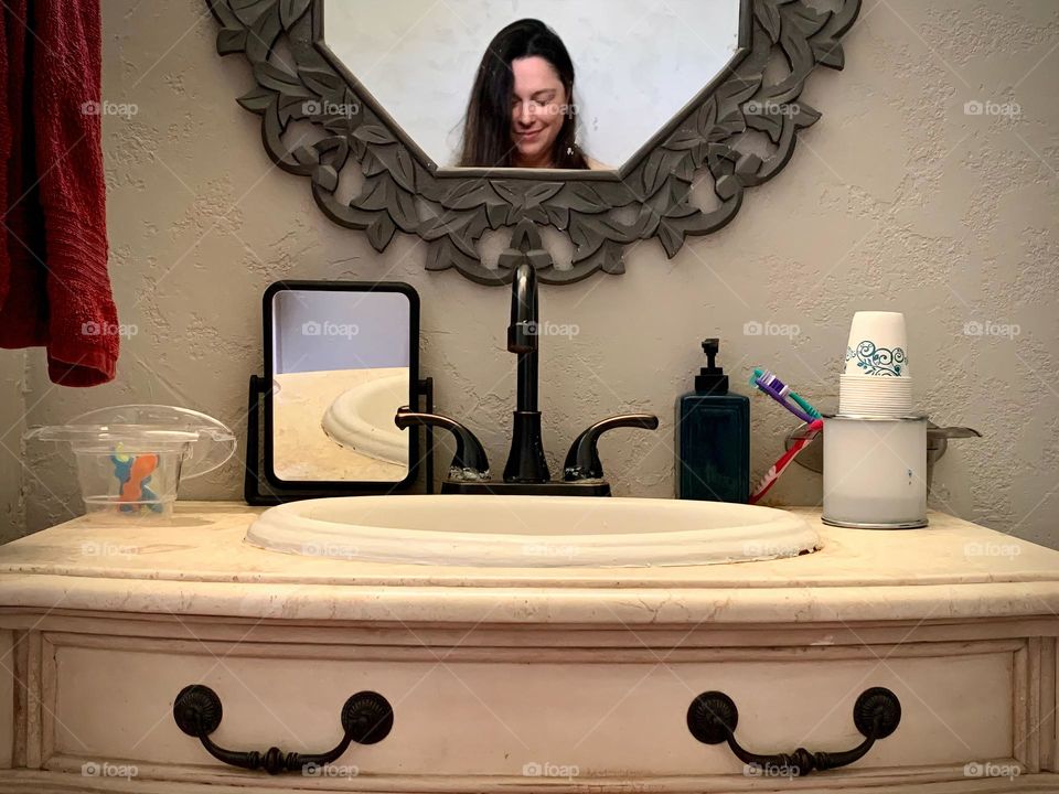 Selfie with mobile phone in the bathroom restroom. Photo seems to see a woman’s head in the mirror and an illusion of the body being the faucet and handles. Symmetry and face looking down.