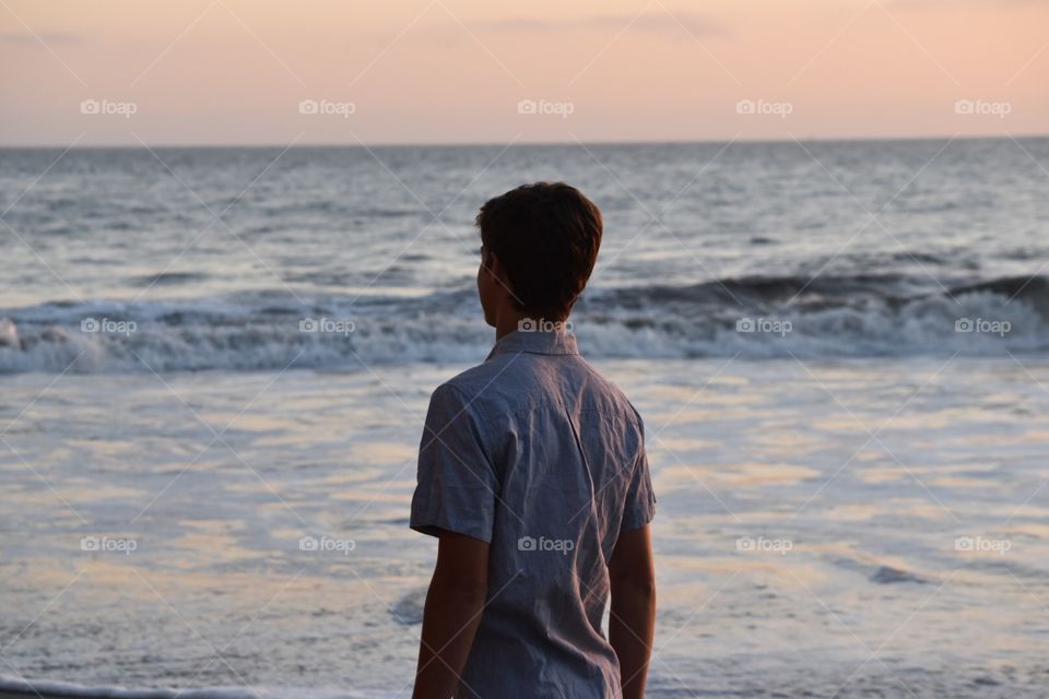 boy with blue collared shirt looking at ocean with orange sunset and waves in background 