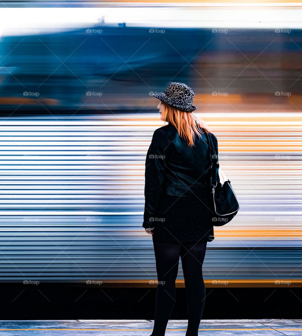 Woman in black with orangey yellow hair stands in front of a metallic and yellow train carriage that whizzes past