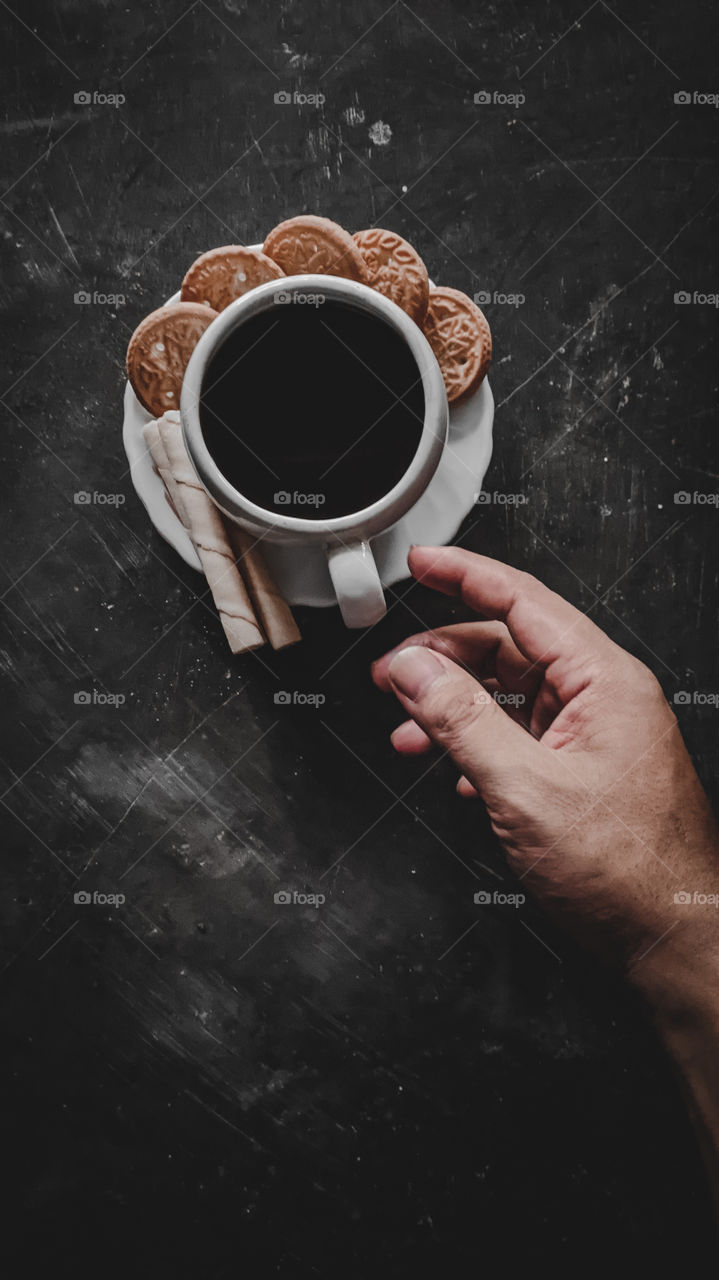 Hands are reaching for a cup of black coffee with biscuits on the floor
