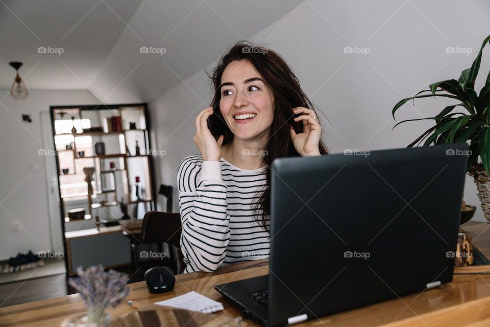 Young woman smiling, while talking on the phone, sitting at her desk office, with a laptop in front.