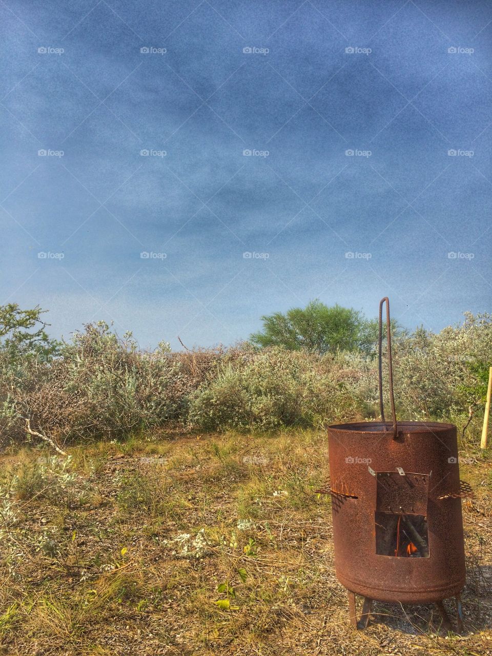 A rusty little barrel with a small fire within sits in a grassy clearing. Desert shrubs lie behind it. A blue sky is overhead with some wispy clouds spread throughout it. 