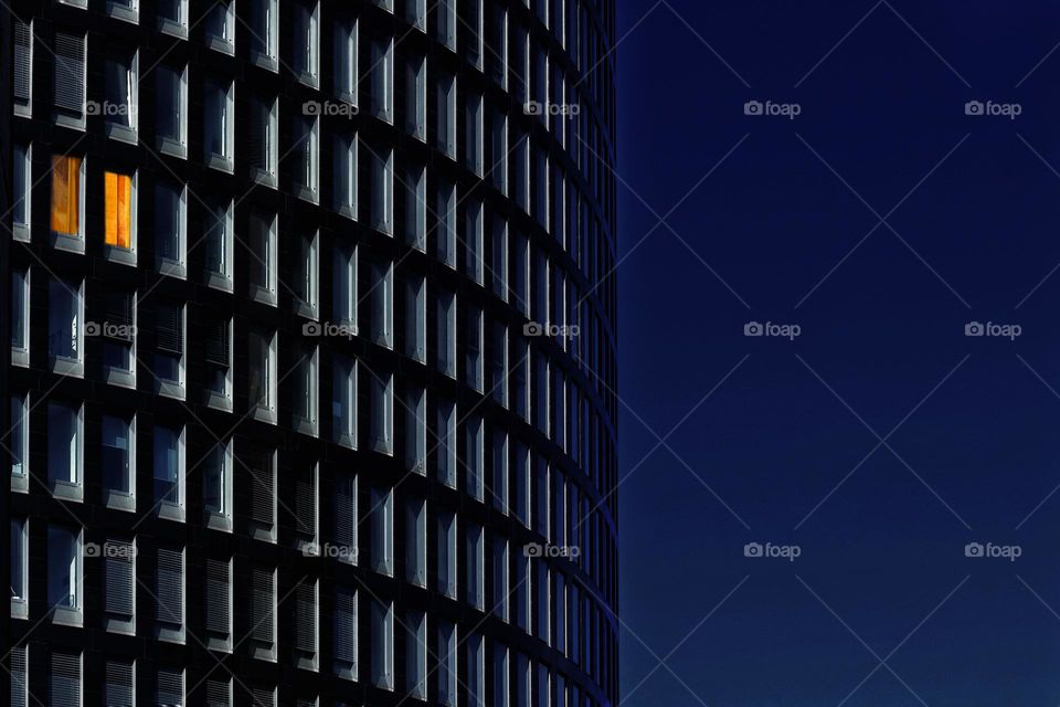 A skyscraper office with many dark windows at blue hour in which golden light shimmers from two windows