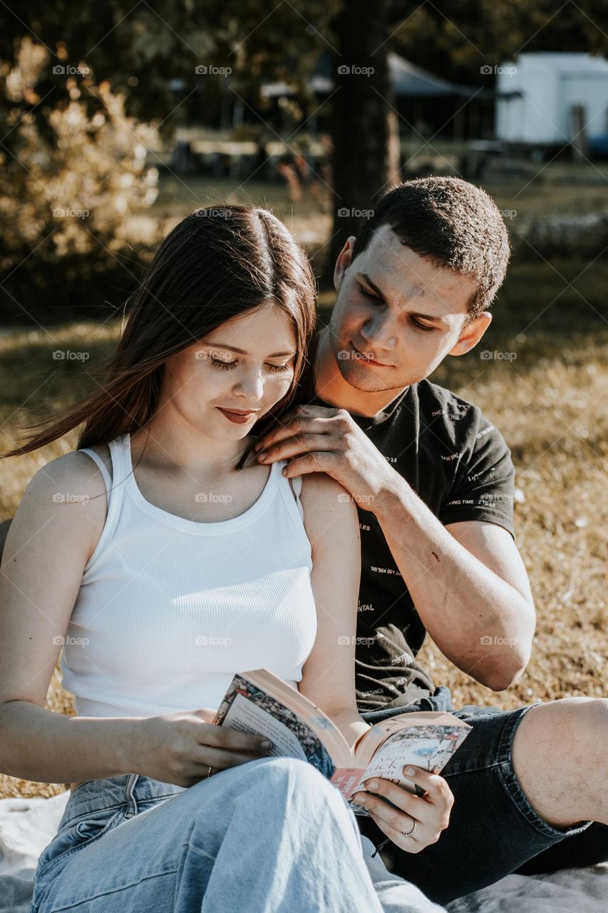 Portrait of one young beautiful Caucasian couple on a picnic: the girl is reading a book, and the guy is straightening her hair while sitting on a blanket in the park on a summer day, close-up side view.