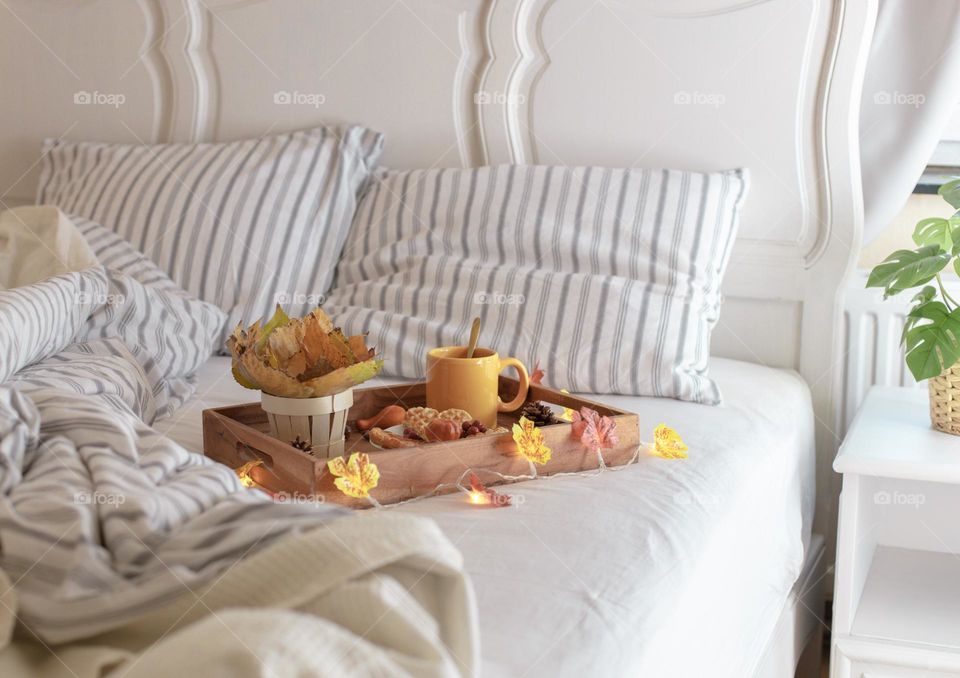 Brown wooden tray with breakfast waffles, a mug of tea, a bouquet of autumn poplar leaves in a basket and a burning garland lie on a carpet with gray striped linens in the bedroom, side view cope plan. The concept of breakfast in bed.