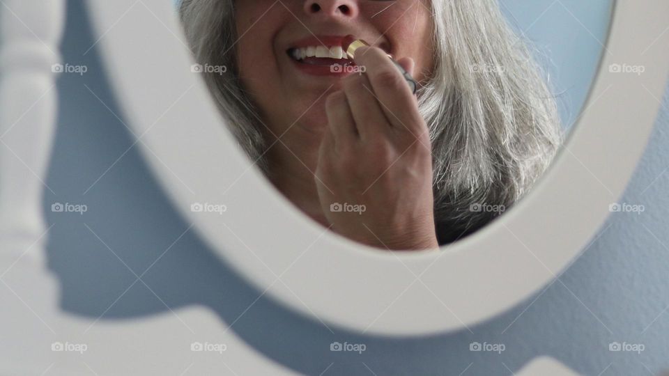 Cropped closeup of gray haired young woman putting on red lipstick using mirror on clean white frame and blue background