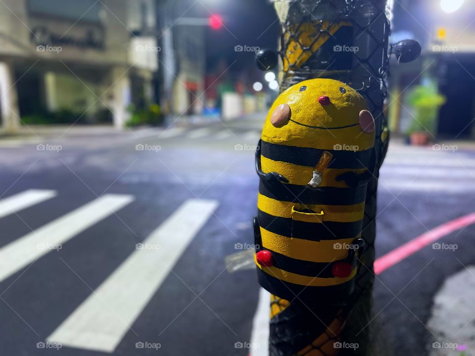 A cigarette garbage shaped like a bumble bee. It is nighttime, and there are stores and a crosswalk in the background. 