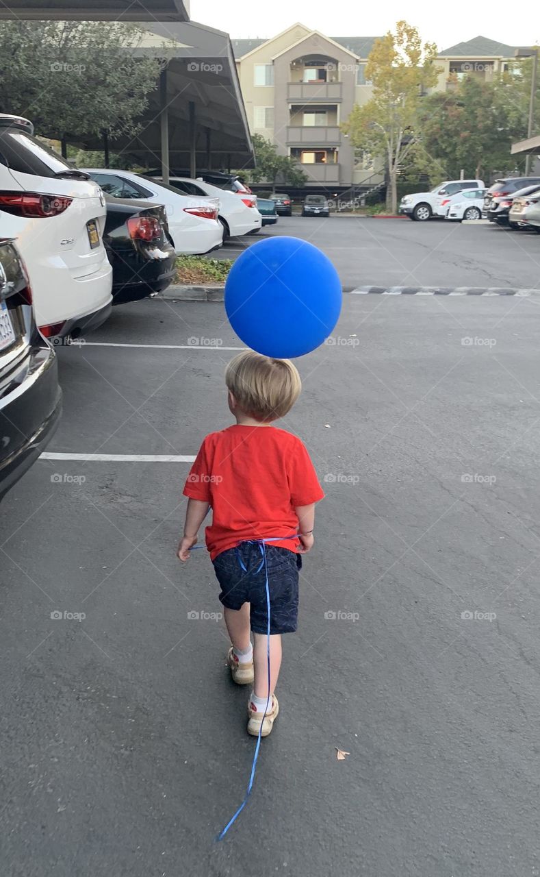 Toddler boy in a vibrant and bright red shirt with his round blue balloon celebrating a birthday from a party.  He’s walking along the cars in the apartment parking lot