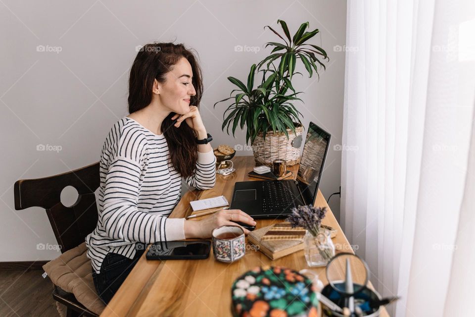 Young woman smiling, while working at her desk, using a laptop, surrounded by different objects.