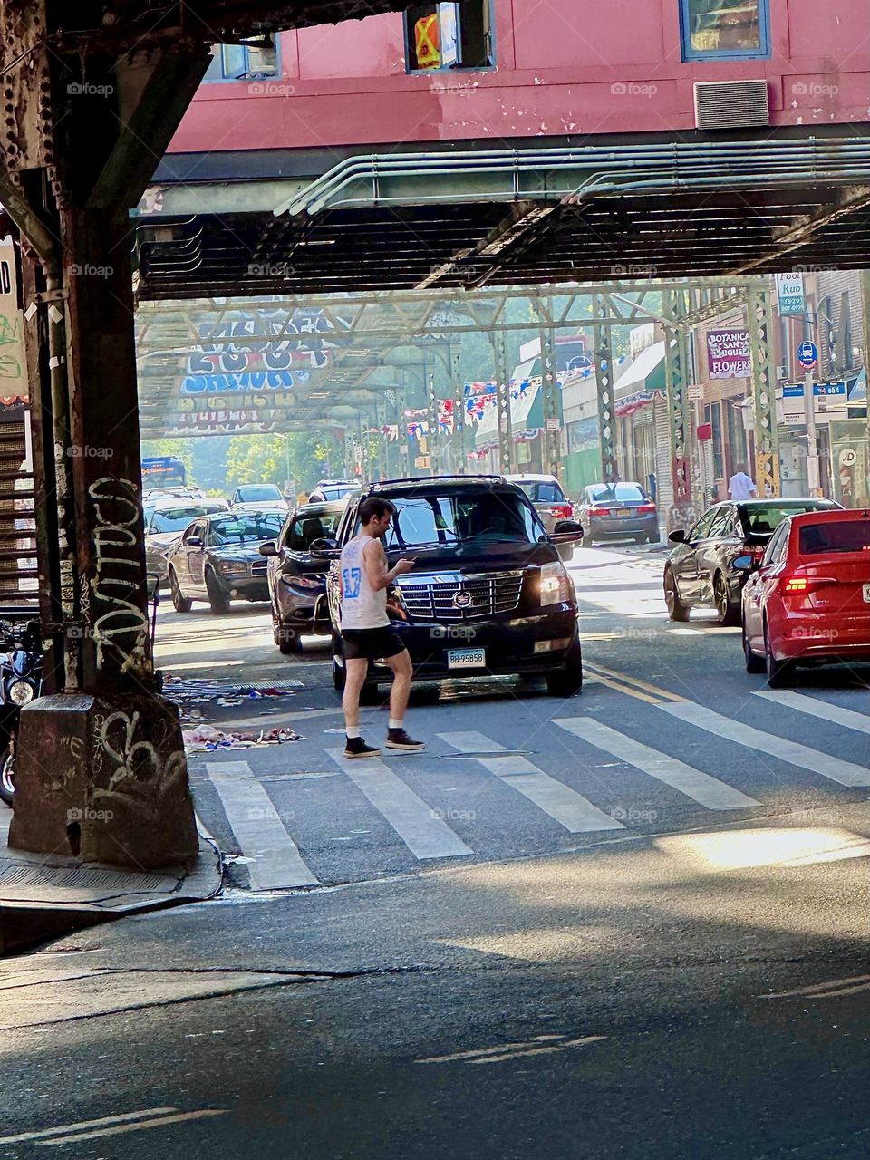 A pedestrian is crossing on “Broadway” at the busy intersection of “Jefferson St” in “Bushwick”, Brooklyn underneath the overpass of the “M” train at the “Myrtle Ave” station. 2024. Hypnotic Productions