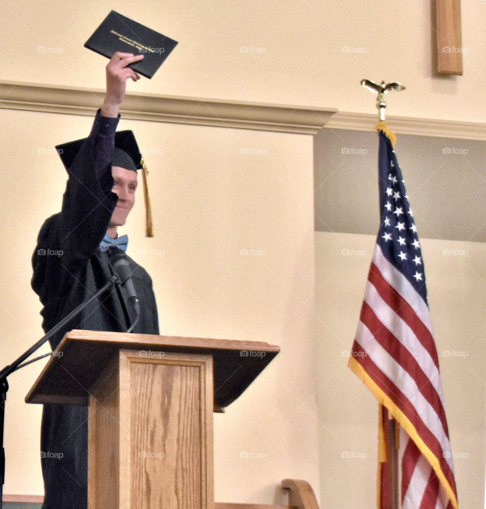 Graduate wearing cap and gown, holding up diploma. Standing by the American flag