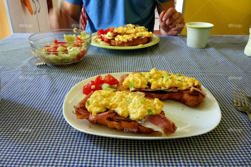 Scrambled eggs on a croissant with bacon and leek on a withe plate with tomato and cucumber salad