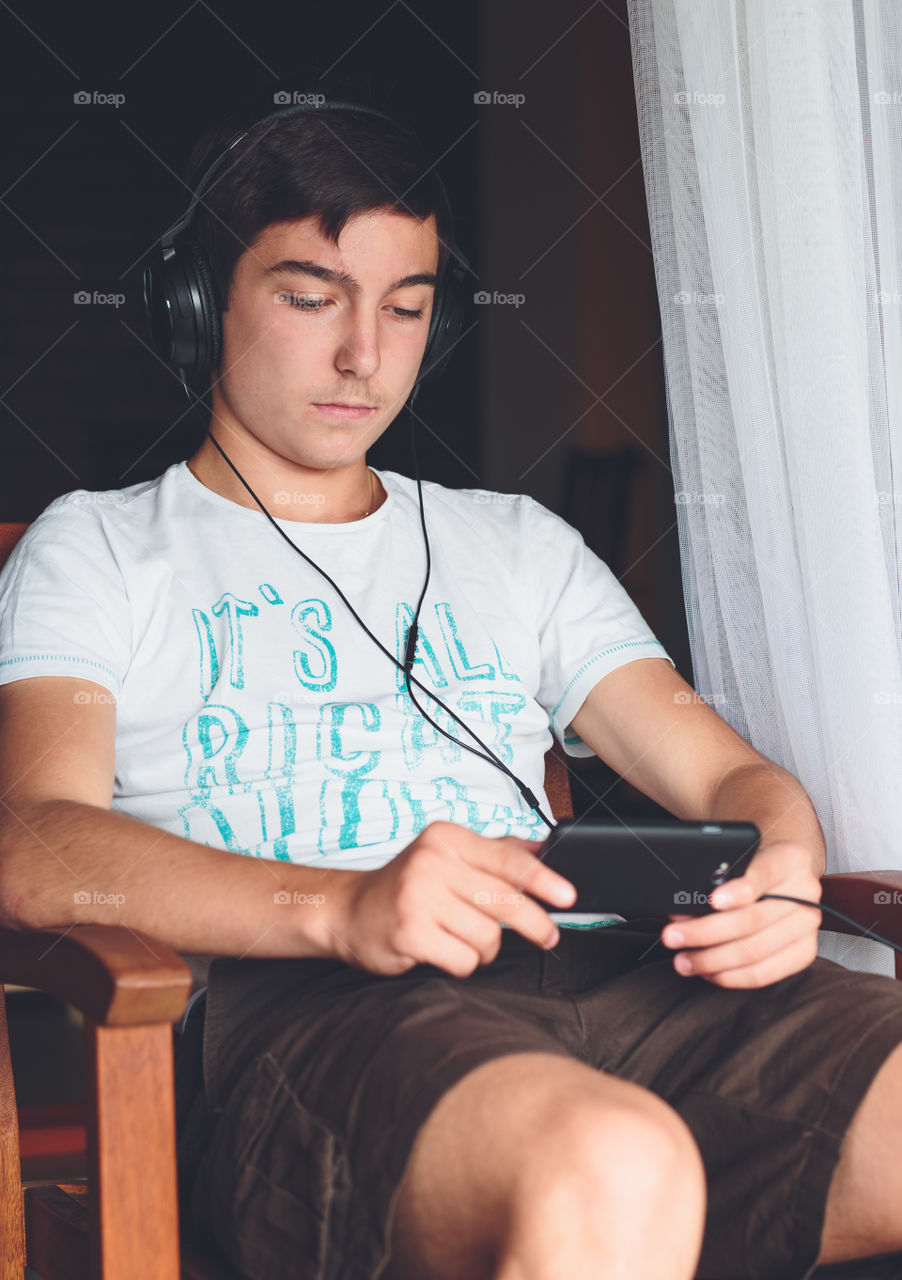 Concentrated on mobile phone. Young boy sitting at the patio door and listening to music from mobile phone