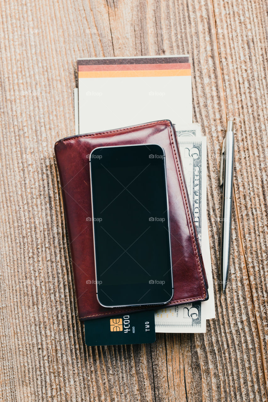 Smartphone with blank screen, wallet, dollar banknotes, debit credit cards and notebook on wooden table. View from above. Portrait orientation