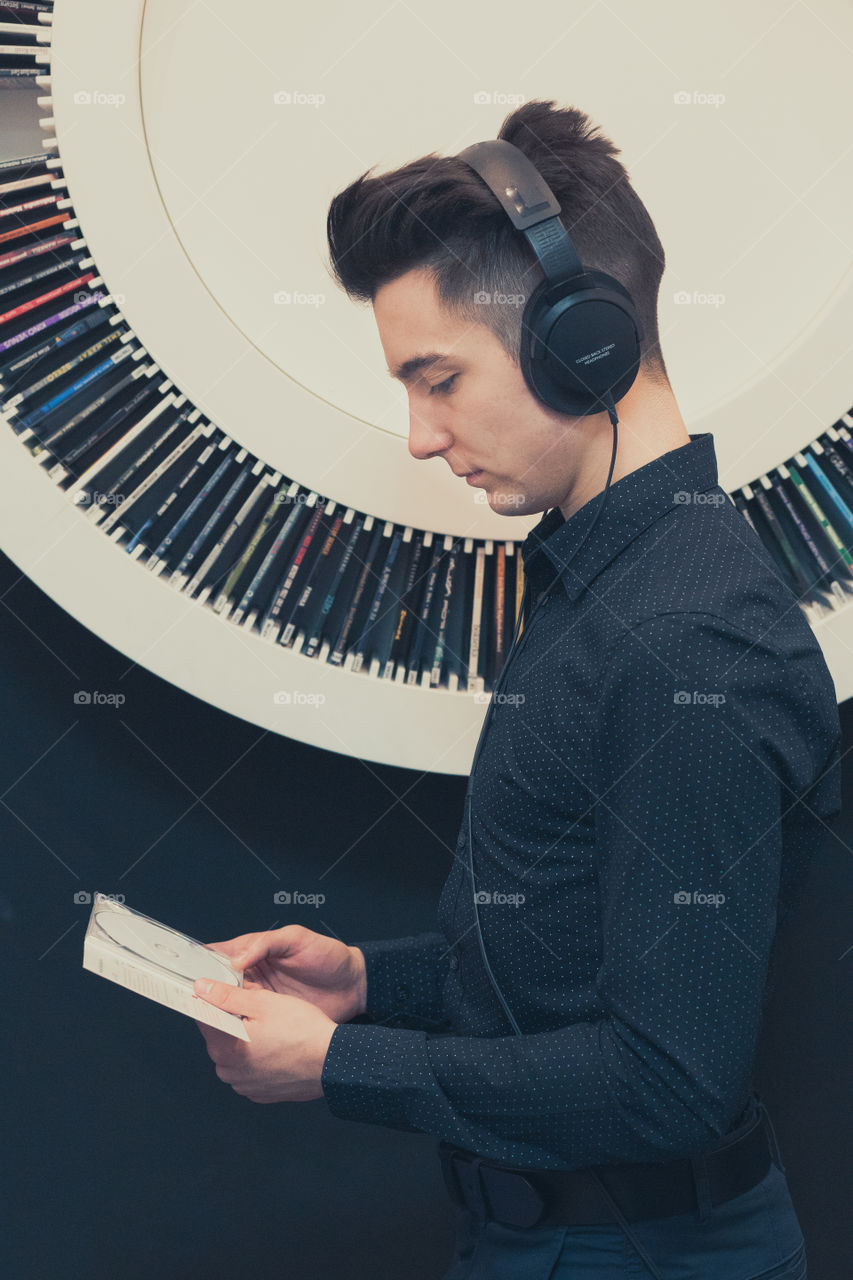 Young man listening to music through headphones standing next to bookshelf with recordings