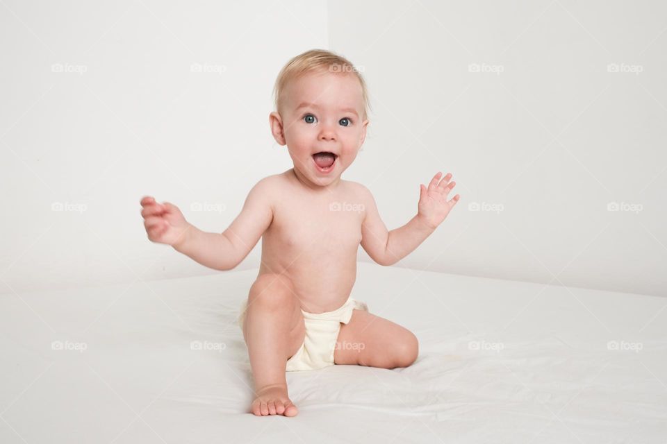 Happy smiling baby sitting on white bed in white bedroom