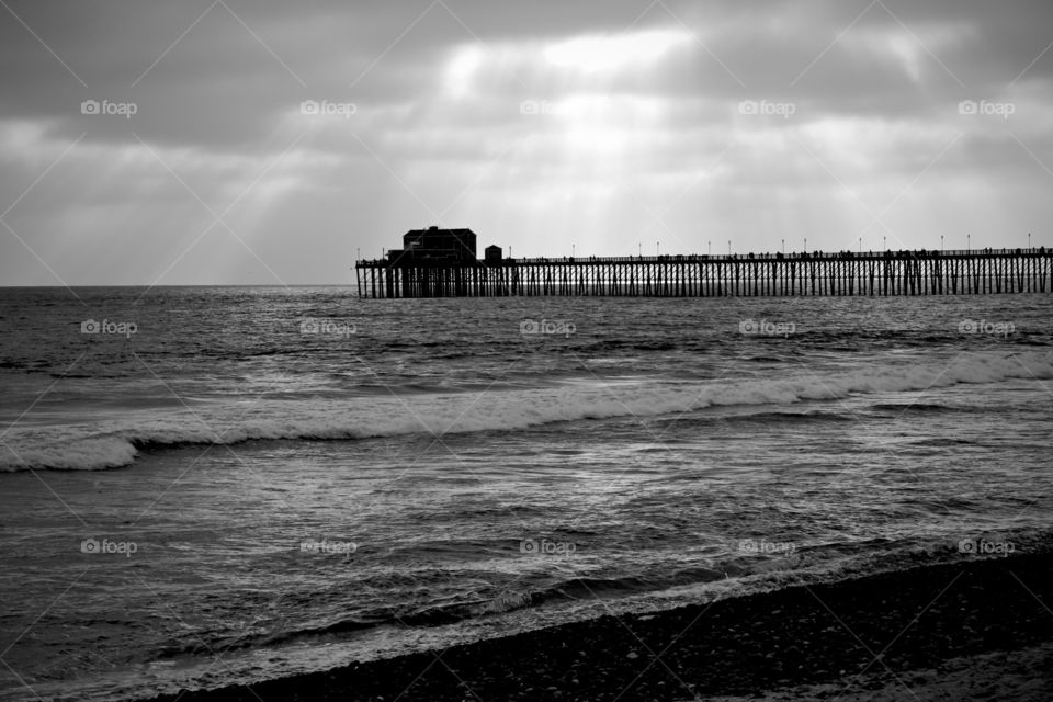 Black and white scene looking out to the pier as the sun starts to come through the clouds. 