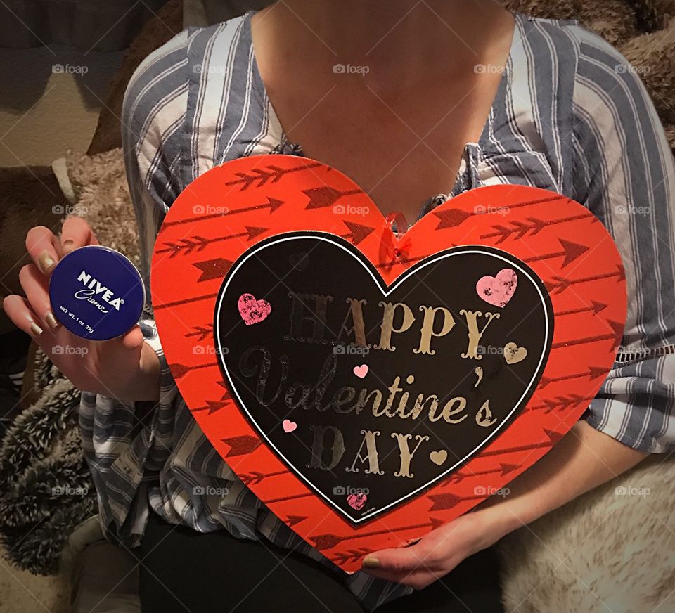 A woman sitting on her bed holding a red heart for Valentine’s Day and a tin can with Nivea cream celebrating Valentines. USA, America