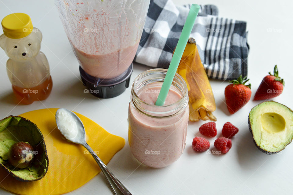 Smoothie and ingredients consisting of banana, raspberry, strawberry, honey and avocado next to a blender