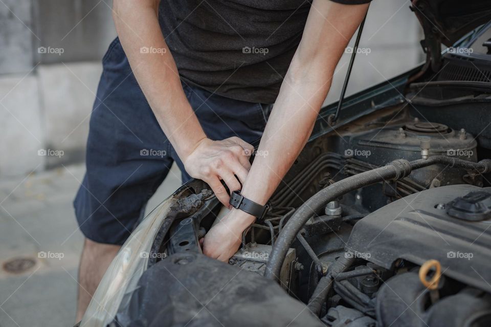 One young Caucasian unrecognizable man pulls out a burnt-out light bulb from the left front headlight while standing stooped on the street during the day, close-up side view. Car repair concept.