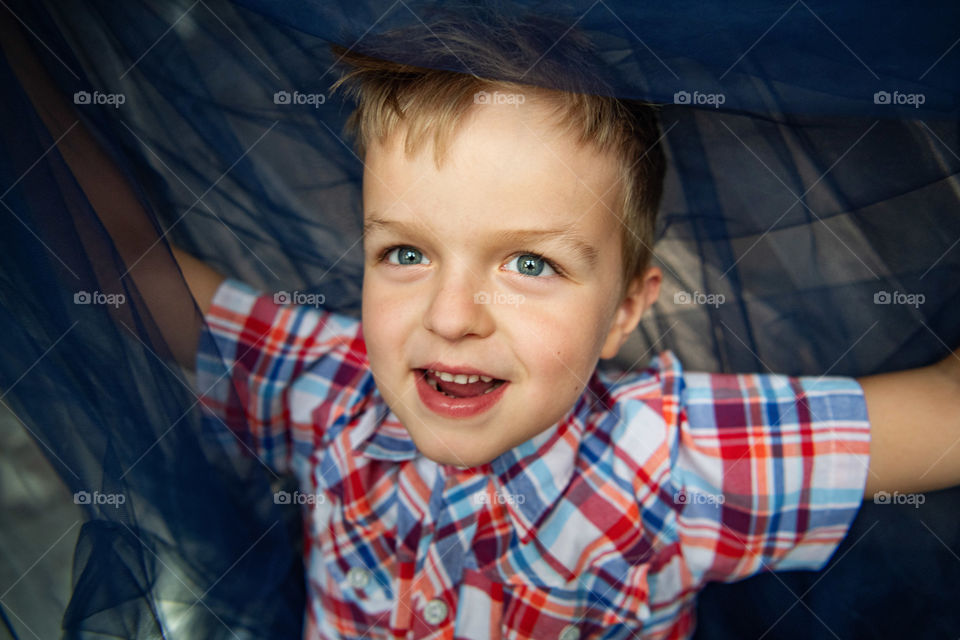 Cheerful blue-eyed boy in a red plaid shirt.  A six-year-old boy peeks out from under a blue bedspread and smiles.