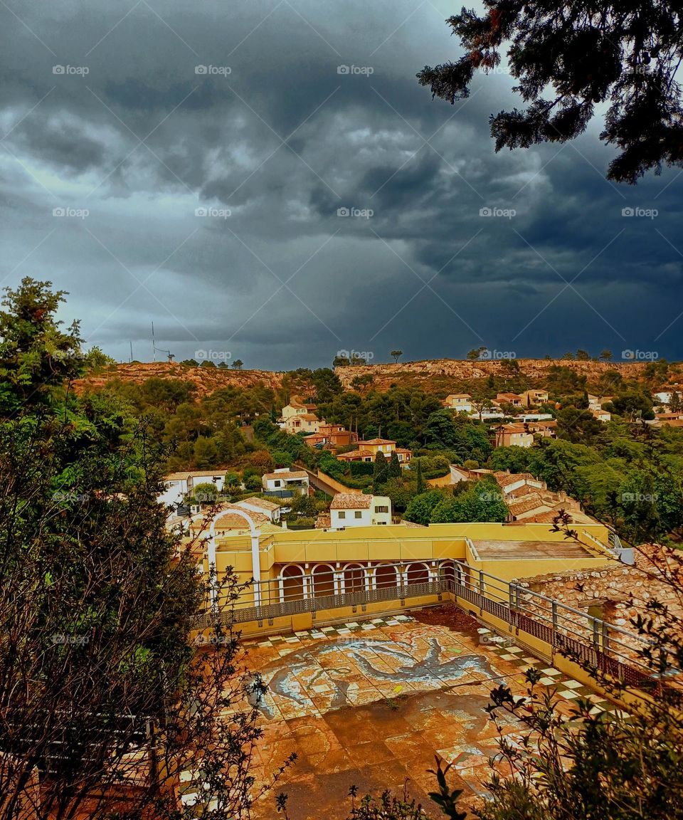 This photo taken in Vitrolles captures a scenic view of a town nestled among lush greenery and hills. The foreground features a patio or terrace with a mosaic-tiled floor, enclosed by a railing.