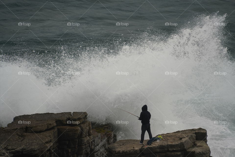 Man fishing off the rocks at the beach as a huge wave breaks around him