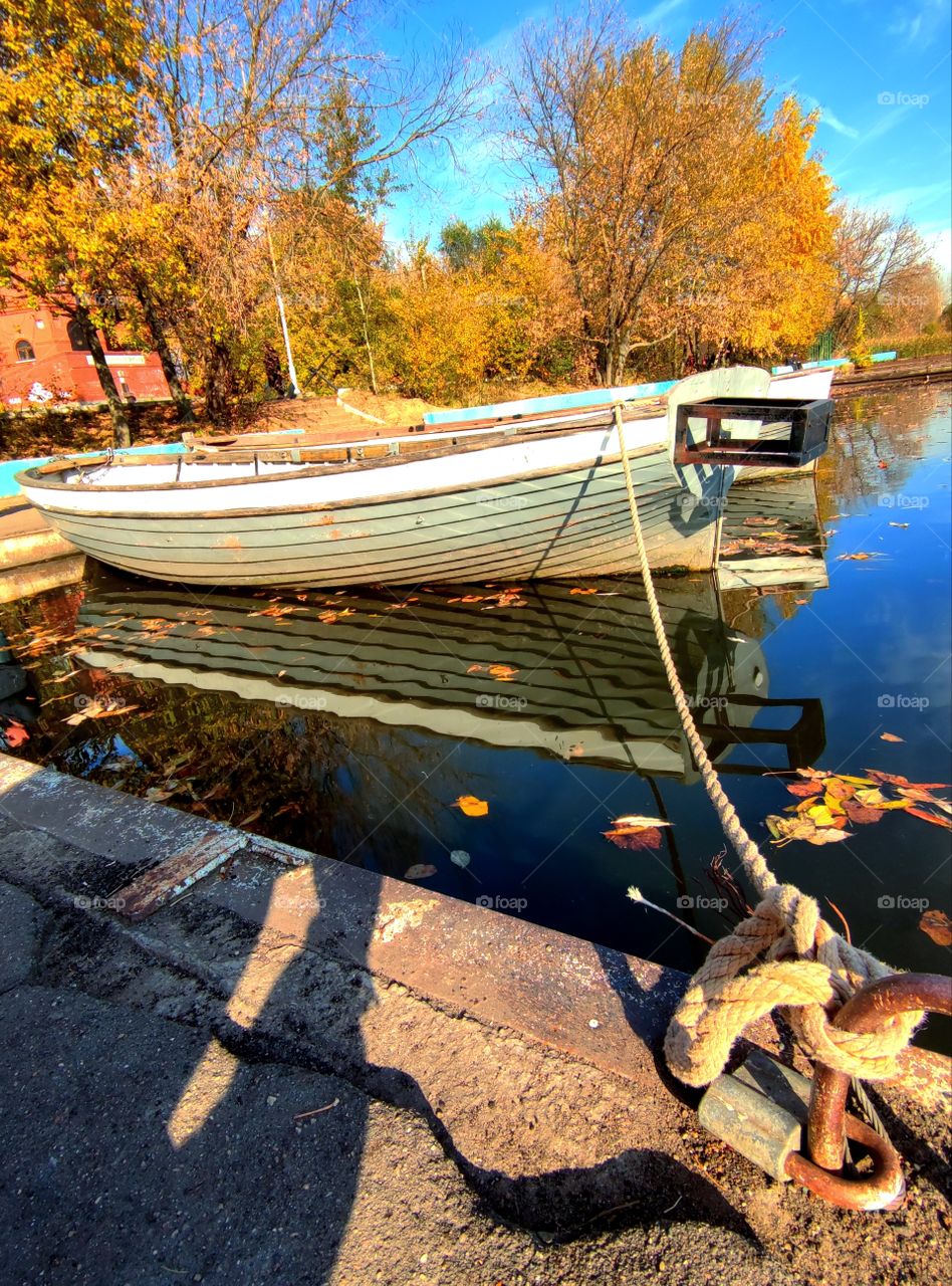 Autumn. Bank of the river. There are yellow trees on the shore. A wooden boat is tied to the pier with a rope. Reflection in the water of the blue sky, boats and autumn trees