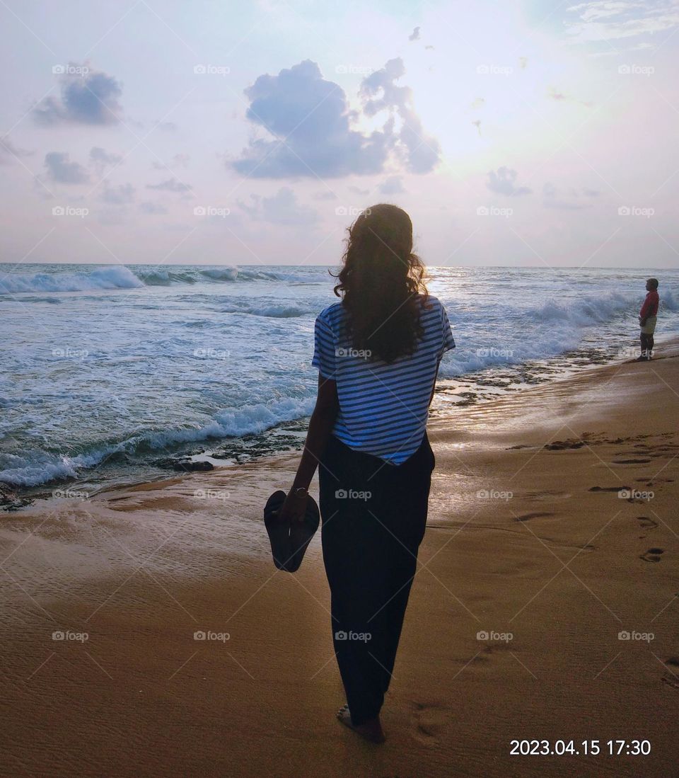 This is a capture of a girl in the sea beach.This is a evening sunsetting scene and the girl is wearing a white colour blouse and a black colour denim.There is a pair of slippers on her hand.There is crashing waves and cloudy sky in the background.