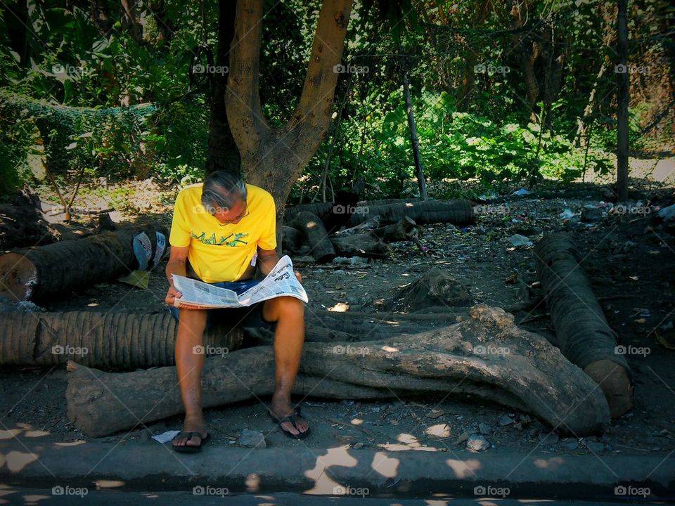 asian man reading newspaper under shade of tree