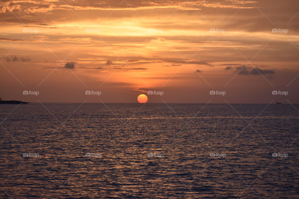 A photo where the sunset begins to slip into the sea horizon. There is a yellow and orange cloudy sky above and below a rippling sea illuminated by the sun itself. Taken in the Carribean Island of Nassau.