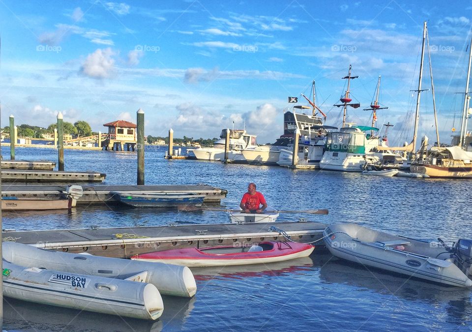 Row row row your boat. A man rowing in a boat next to ships in a marina 