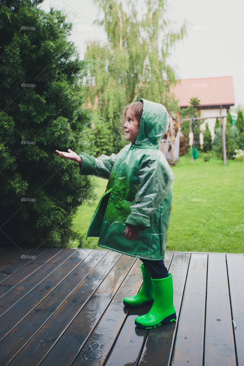 Happy smiling little girl standing on a porch catching raindrops wearing green raincoat