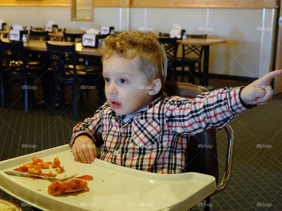 A little boy sitting in a high chair digs into his favorite snack of pepperoni pizza with great vigor. 