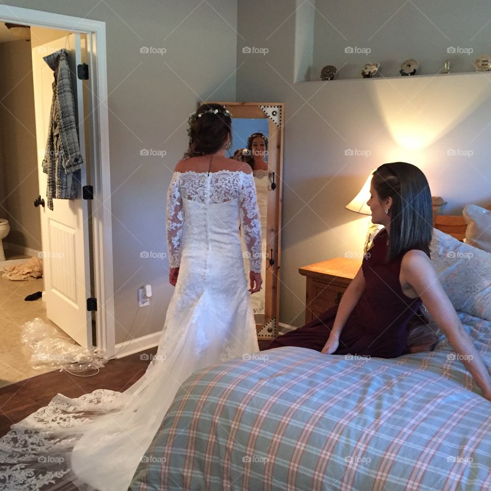 Bride checking her reflection in the mirror on her wedding day as her bridesmaid sits looking on giving her advice.