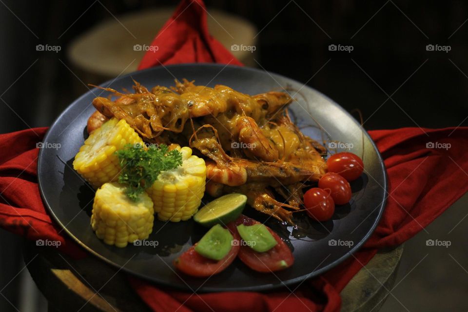 Asian food balado fried prawns with corn chunks, lemon slices and tomatoes served on black plate with red cloth bottom and wooden coaster. Asian style street seafood.