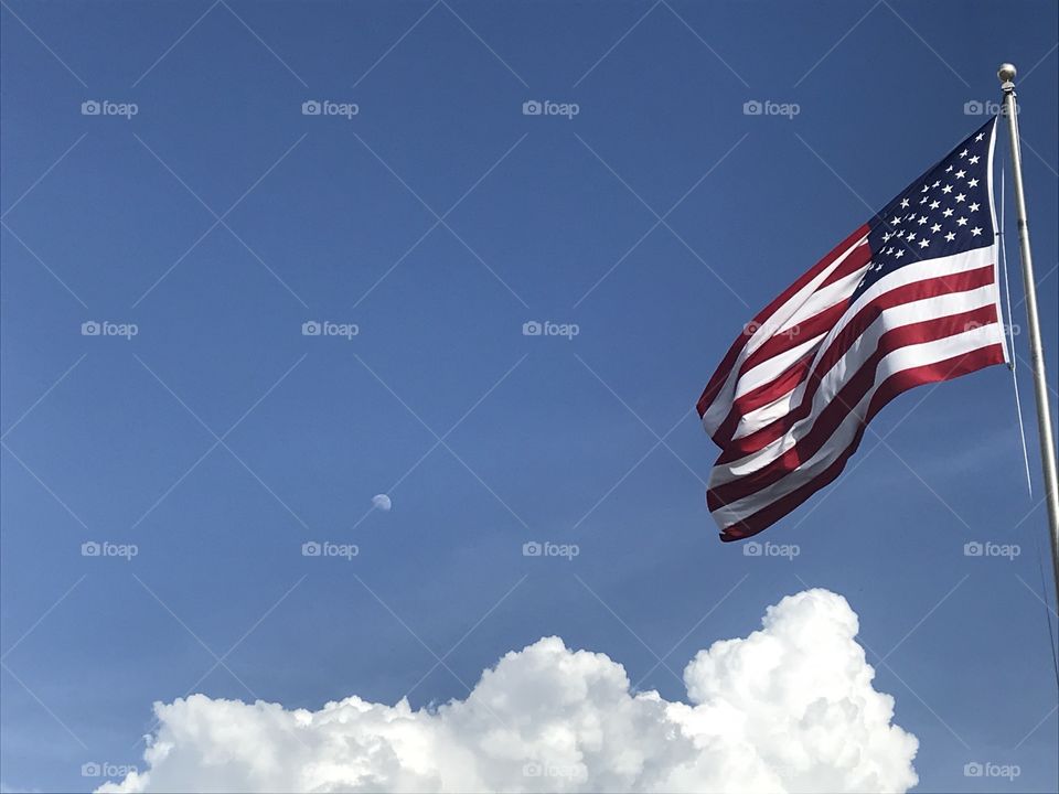 American flag against a blue sky with white puffy clouds. Moon in the distant background 