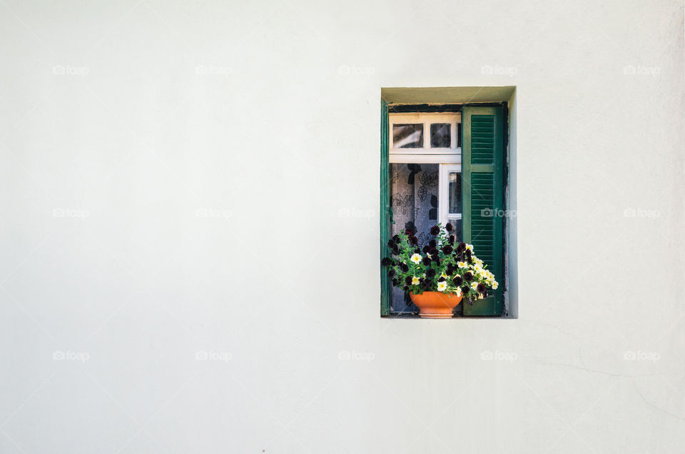 View of the white stone wall of a house and the small green window with curtains and the small pot of colorful flowers.