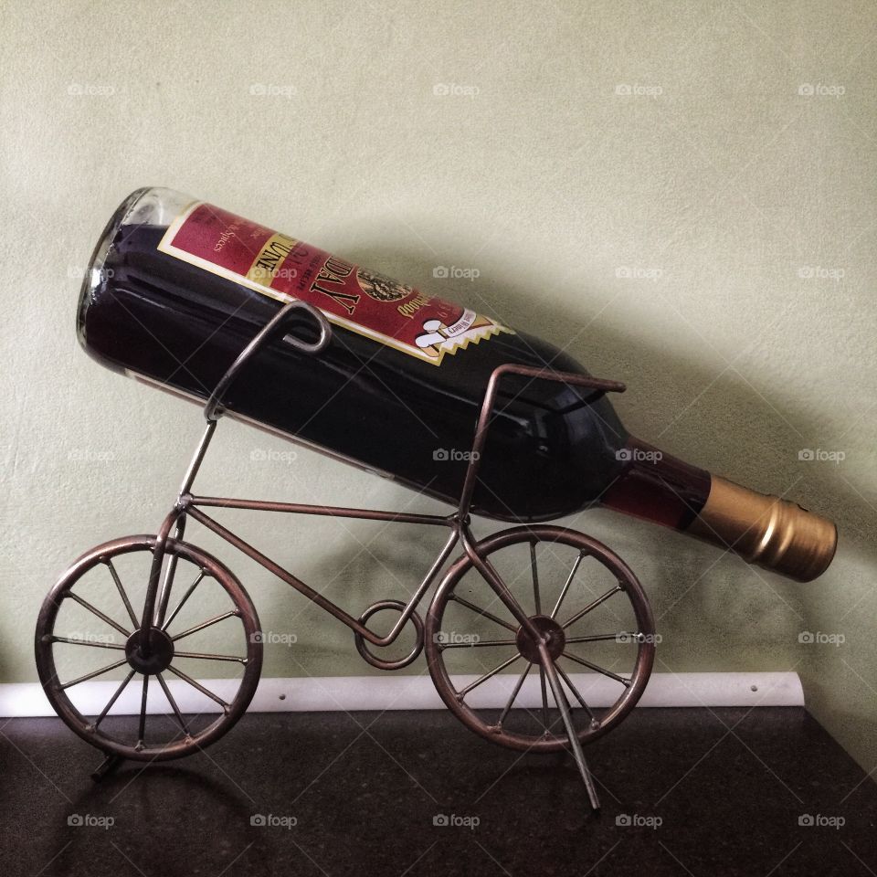 A wine holder shaped like a bicycle holds a full bottle of wine, cork down. It sits in front of a beige wall.