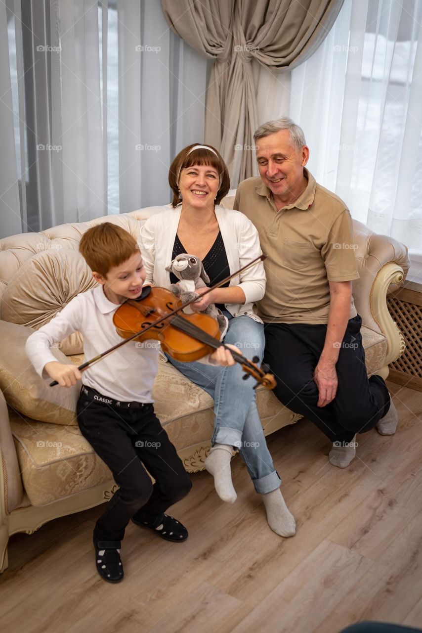 Red-haired boy plays the violin in a bright room, the family listens to music