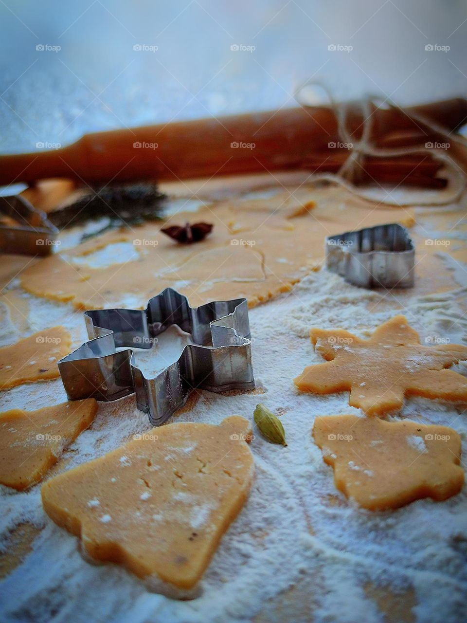 On a board sprinkled with white flour lies a rolled out dough with squeezed out figures. Nearby are figures of their dough, metal molds, a stack of cinnamon sticks, which is tied with twine. In the background lies a wooden rolling pin.